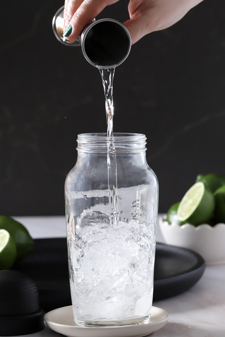 A hand pouring Gin from a jigger into a cocktail shaker half full of ice.