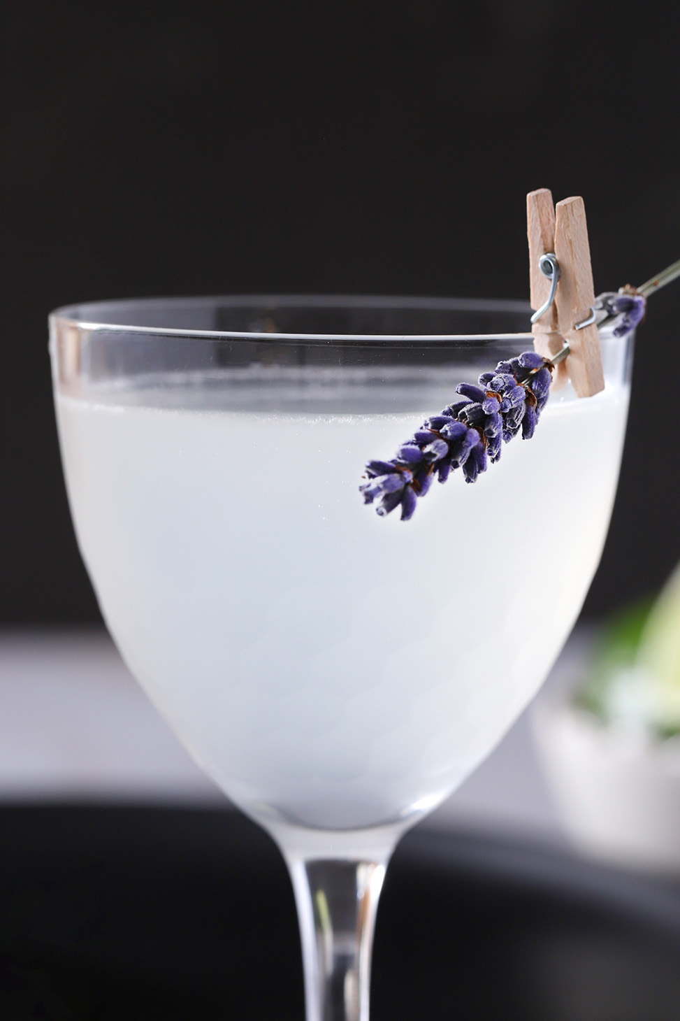 A lavender gimlet garnished with a sprig of lavender clipped to the side of the glass with a tiny clothes pin.