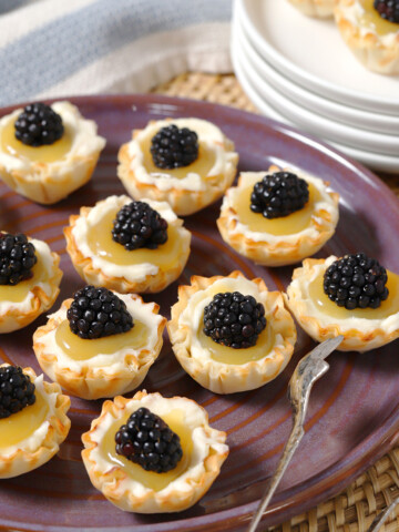 A platter full of mini lemon tarts with blackberries on them.