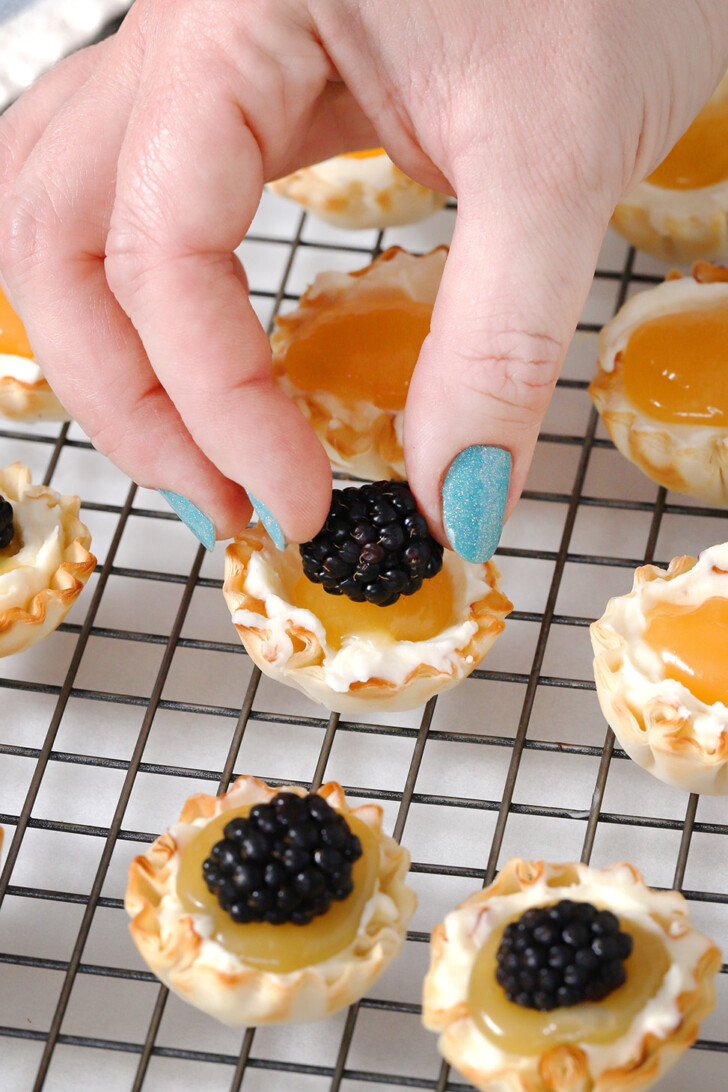 A hand placing a fresh blackberry on a lemon tart.