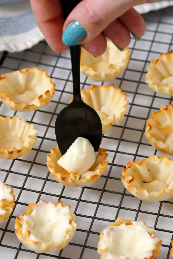 A hand using a black spoon to place a small amount of cheese mixture into a phyllo cup.