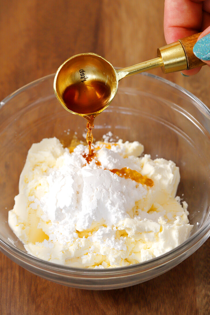A hand pouring vanilla into a glass bowl of mascarpone cheese and powdered sugar.