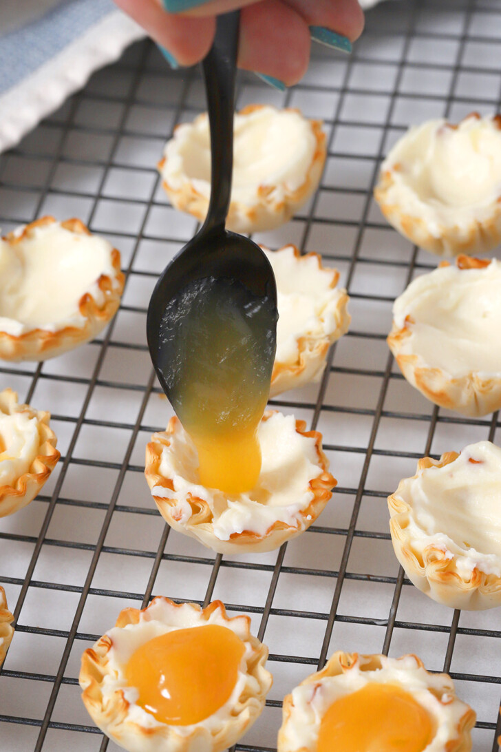 A hand using a black spoon to place a small amount of lemon curd into a phyllo cup.