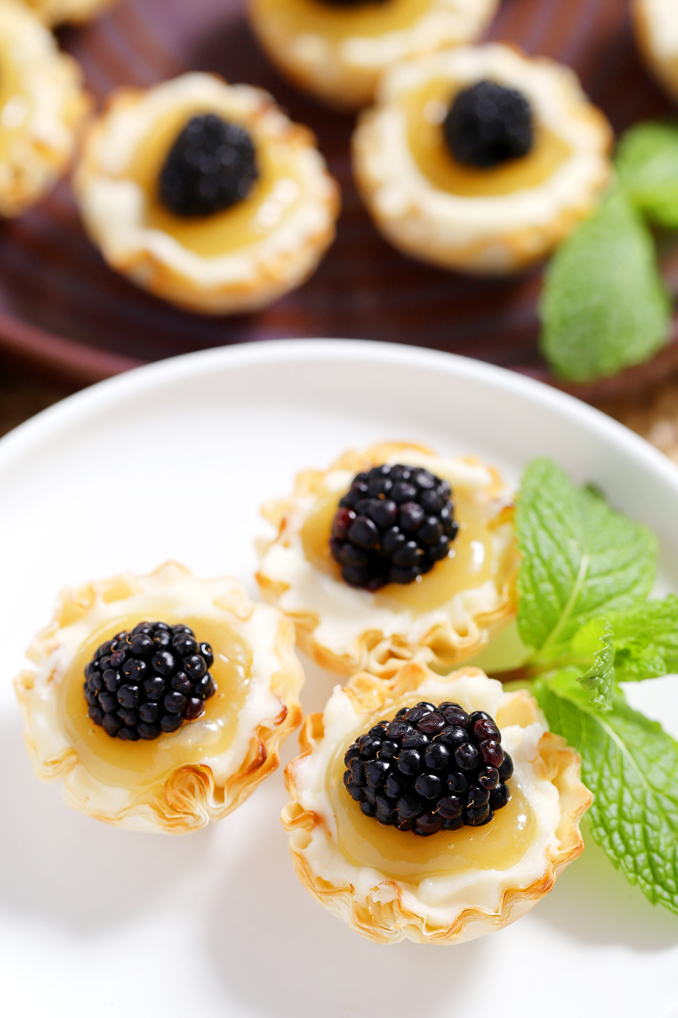 A white plate with 3 mini lemon tarts on it, garnished with mint, with more mini tarts in the background.