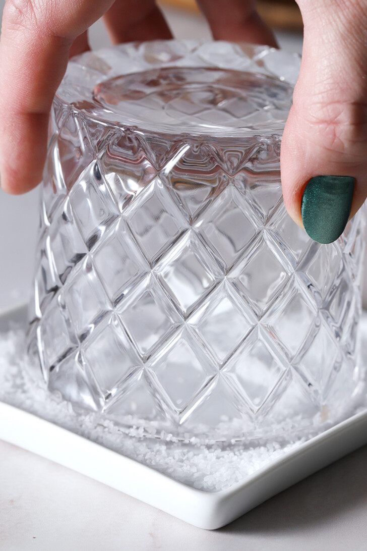 A hand dipping a rocks glass into a small dish full of coarse salt.