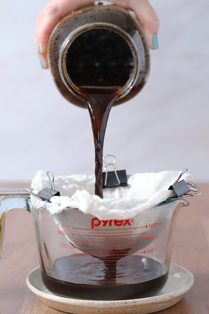 A hand holding a large mason jar and pouring its contents into the cheesecloth lined strainer.