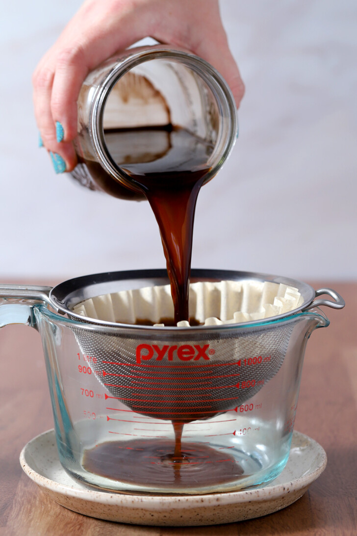 A hand pouring cold brew coffee from a mason jar into a strainer lined with a coffee filter.