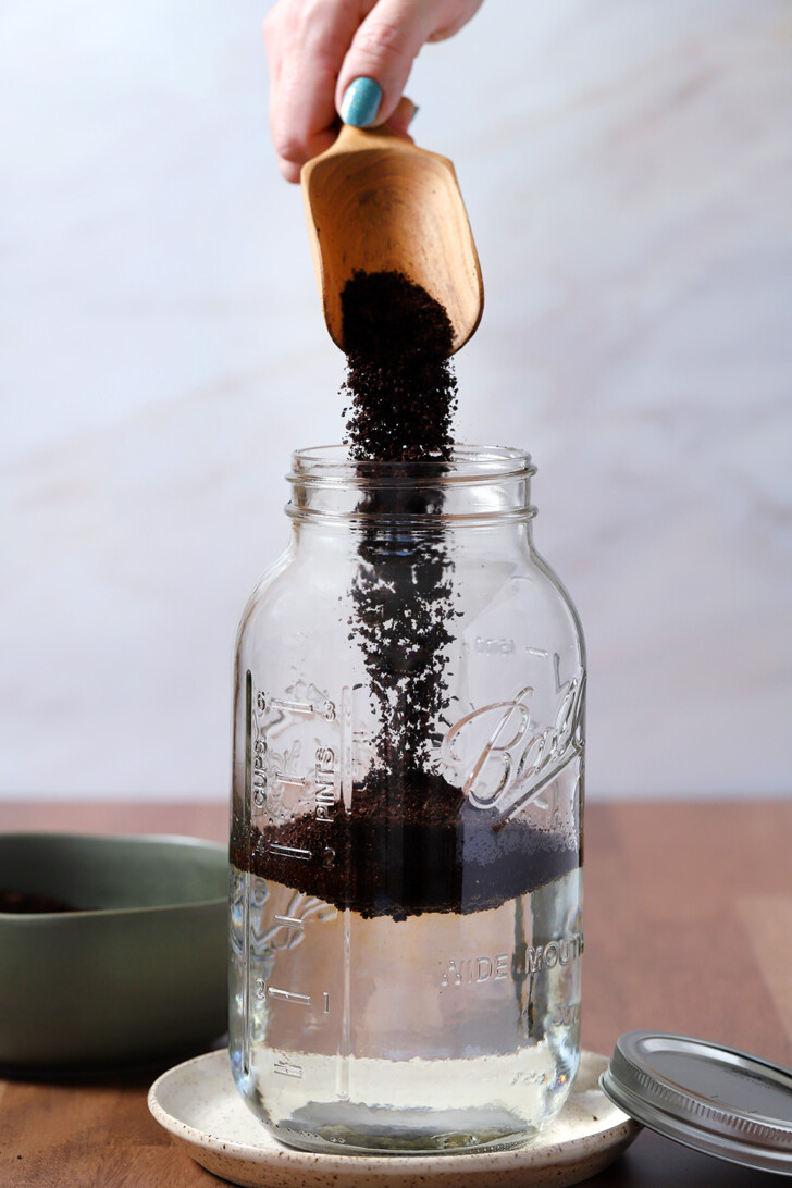 A hand holding a wooden scoop and pouring espresso coffee into a large glass mason jar with water in it.