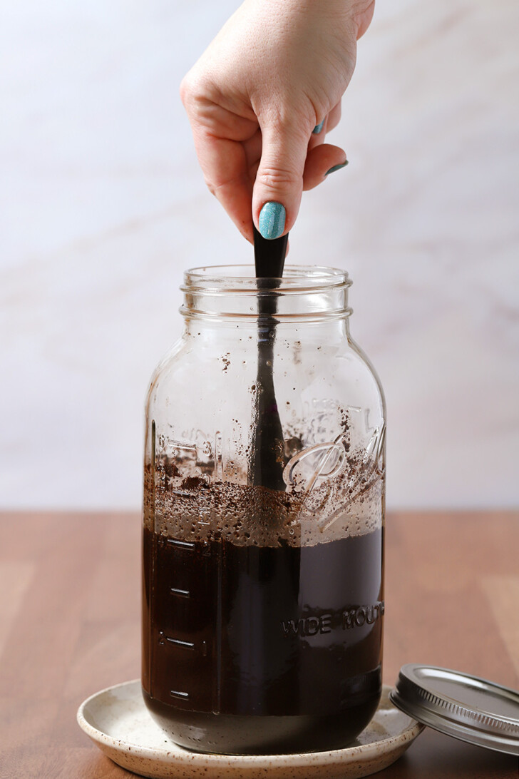 A hand using a butter knife to stir coffee ground into water in a large glass mason jar.