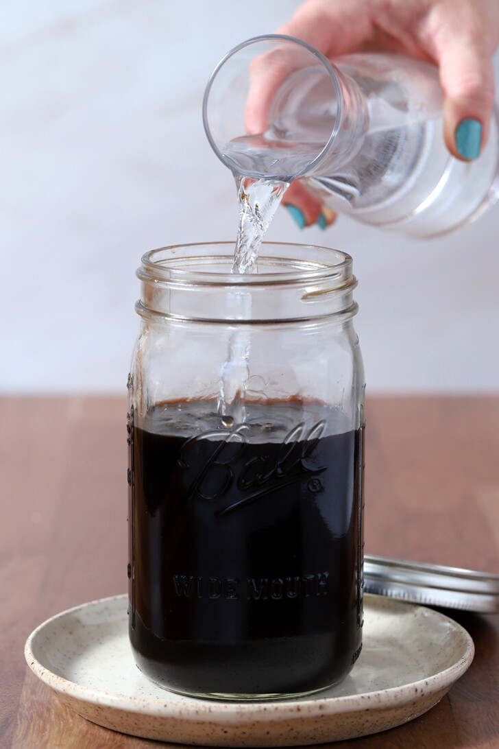A hand pour water from a carafe into a large jar of espresso cold brew coffee.