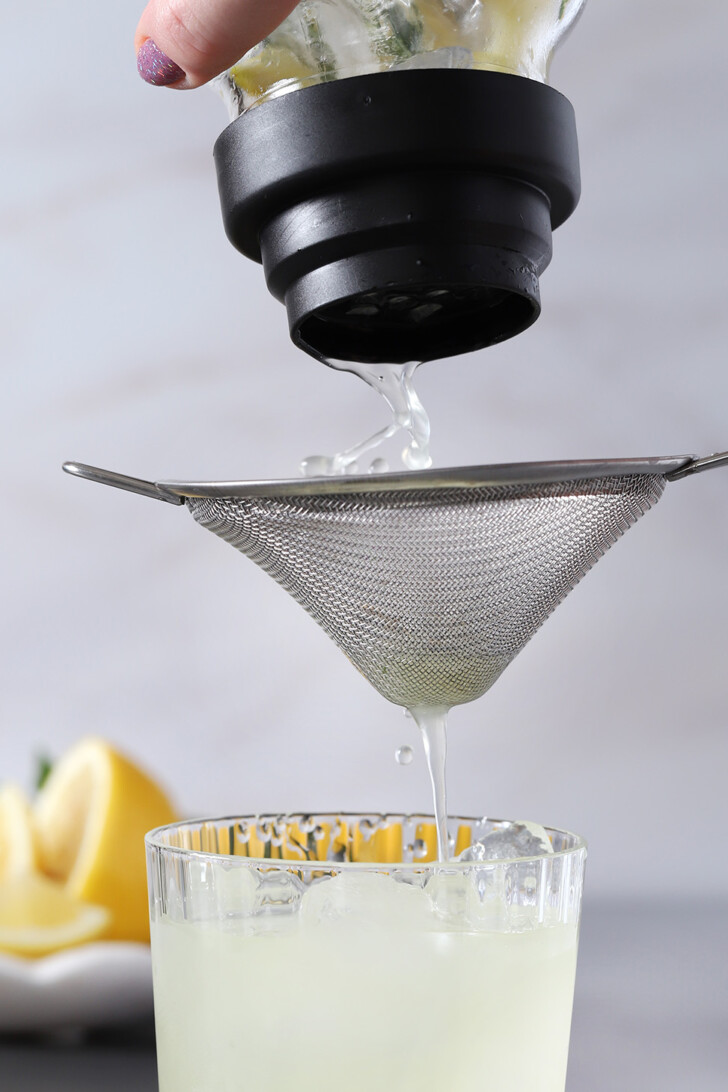 A hand pouring gin smash cocktail from a cocktail shaker, through a fine mesh strainer, into a glass filled with ice.
