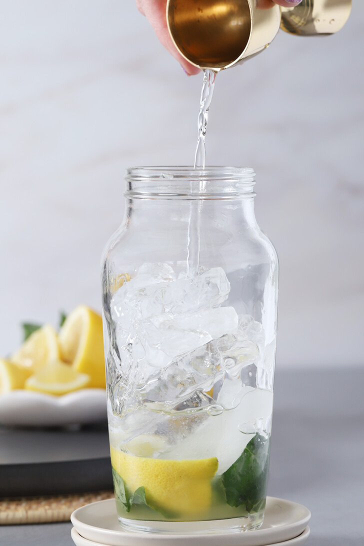 A hand pouring gin into a cocktail shaker with basil, syrup, and lemon in it.