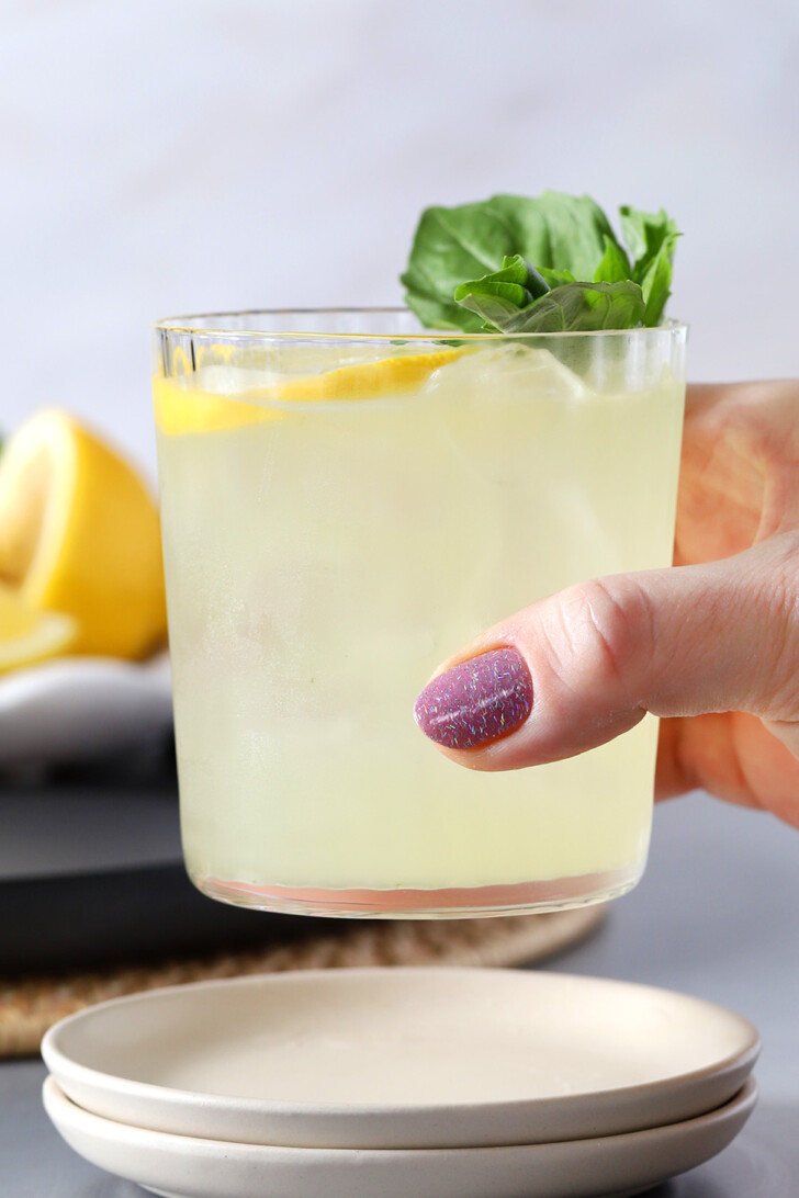 A hand picking up a glass of Gin Smash cocktail garnished with a lemon wheel and fresh basil leaves.
