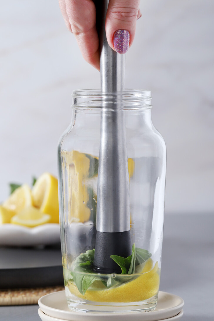 A hand muddling lemon, basil and simple syrup in a cocktail shaker.
