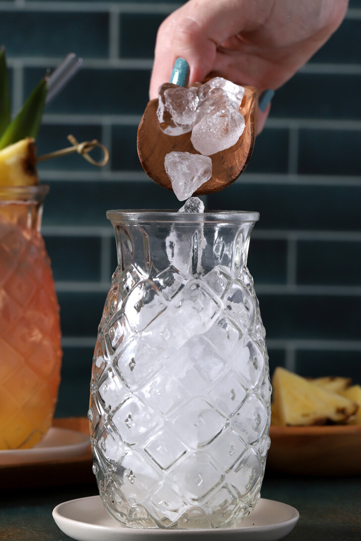 A hand pouring ice from a scoop into a pineapple shaped glass.