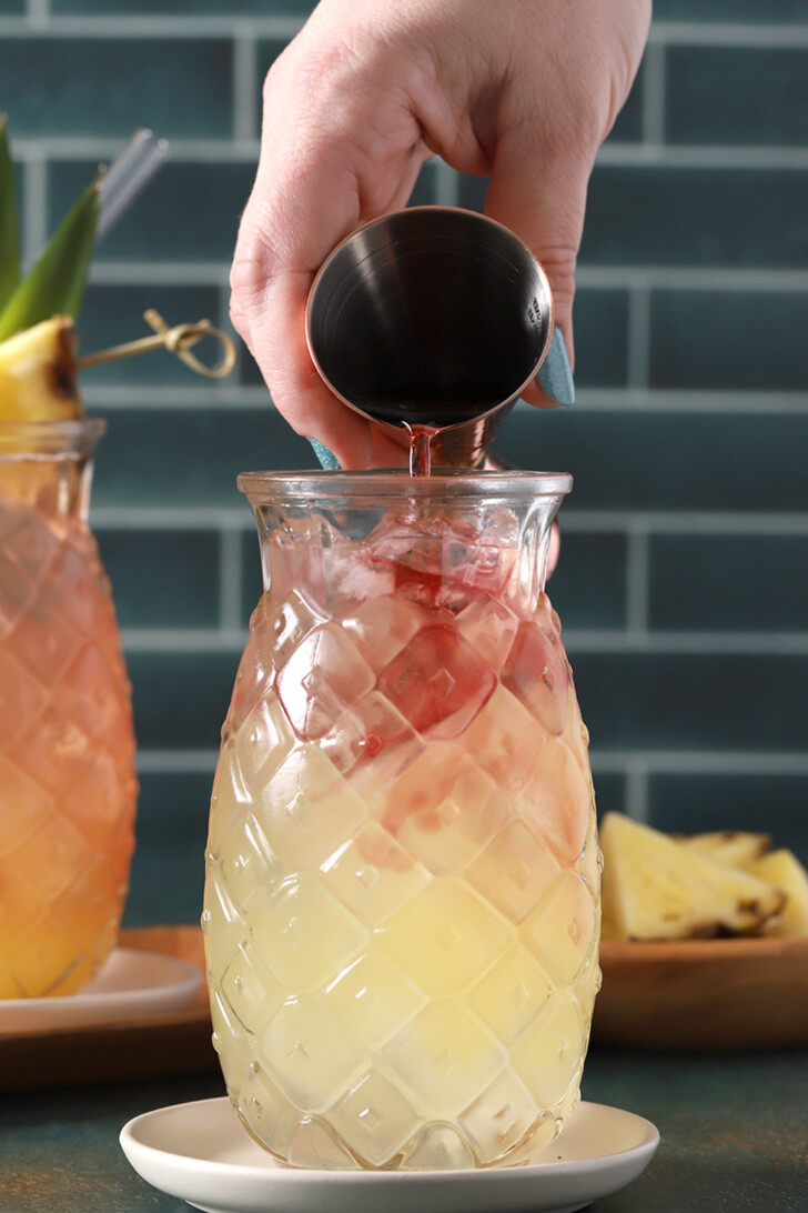 Cranberry juice being poured into a pineapple shaped glass filled with Malibu rum, ice and pineapple juice.