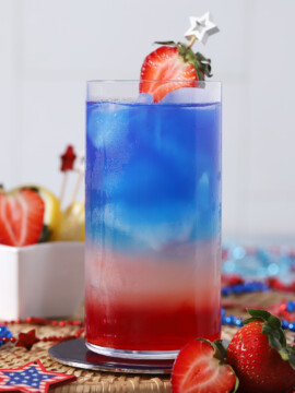 A glass full of red, white, and blue drink layers, garnished with half of a strawberry.