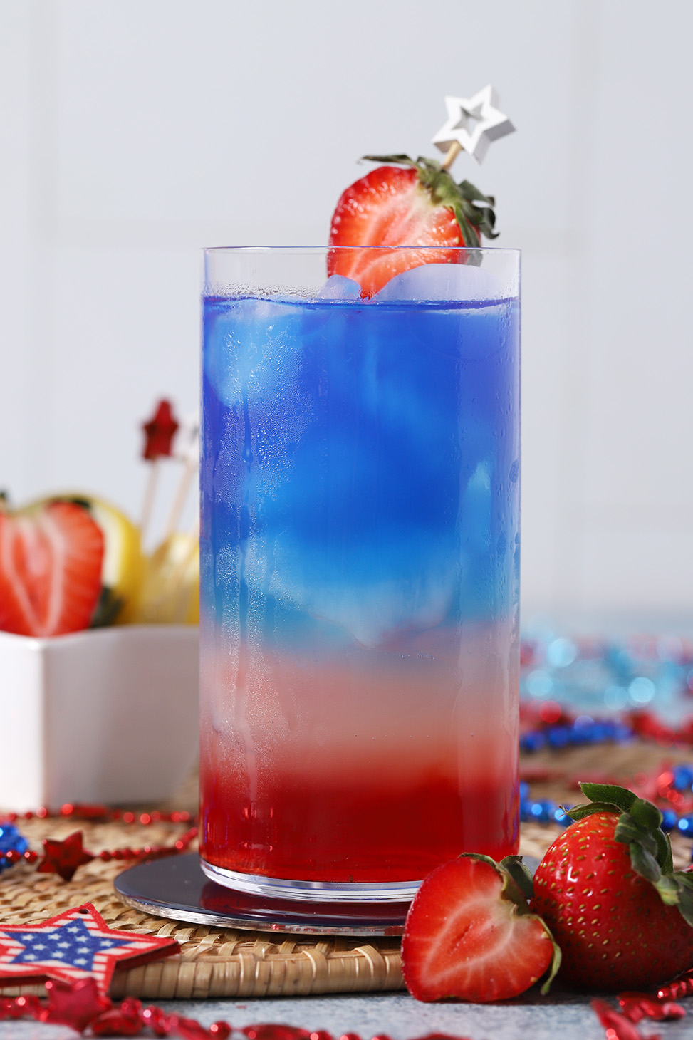 A glass full of red, white, and blue drink layers, garnished with half of a strawberry.