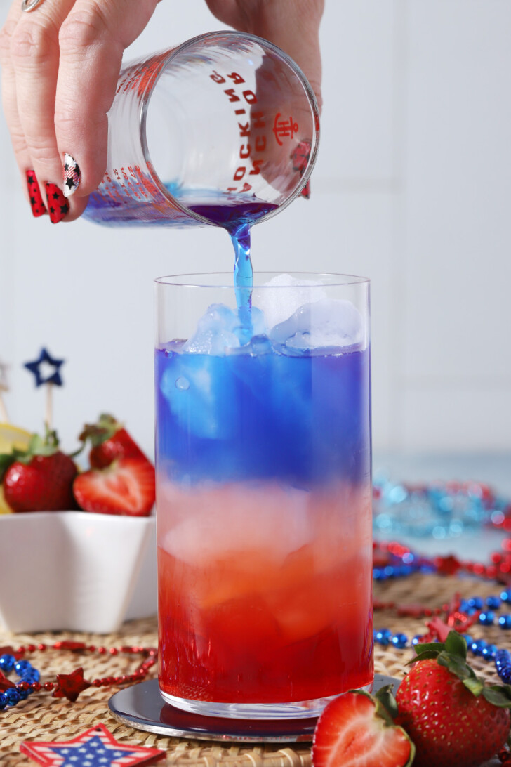 A hand pouring blue water from a measuring glass into a tall glass.
