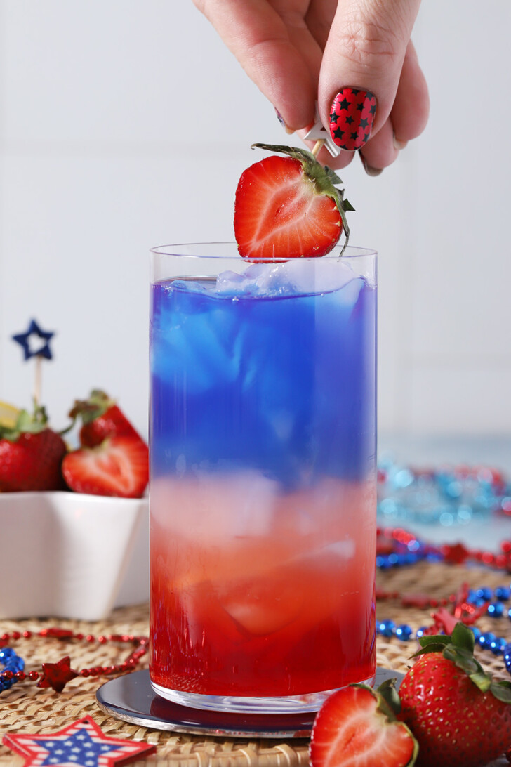 A hand placing a strawberry garnish on a red, white and blue mocktail.