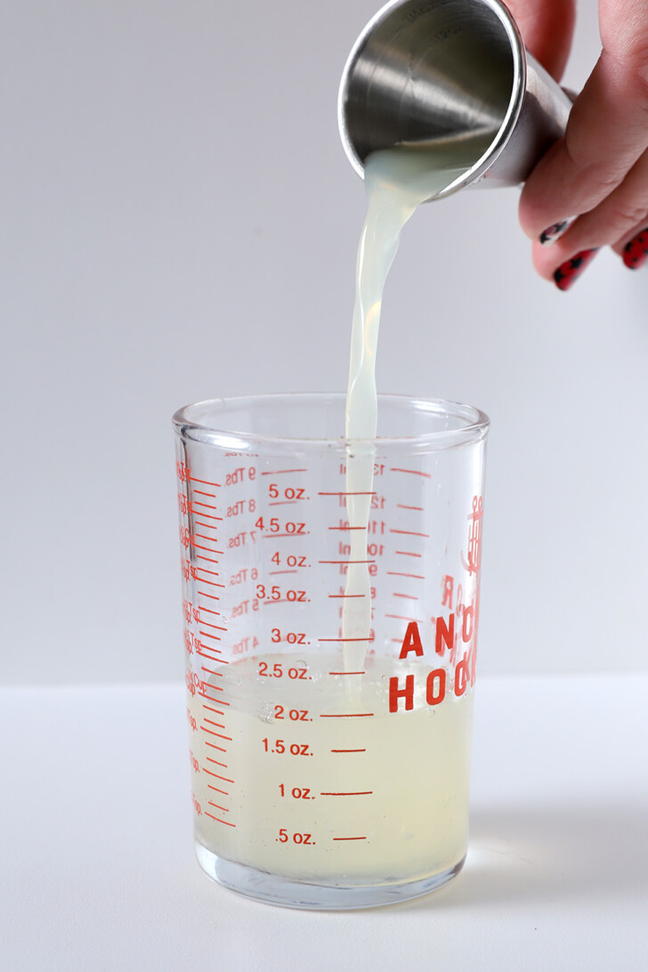 A hand pouring lemon juice from a jigger into a measuring glass.