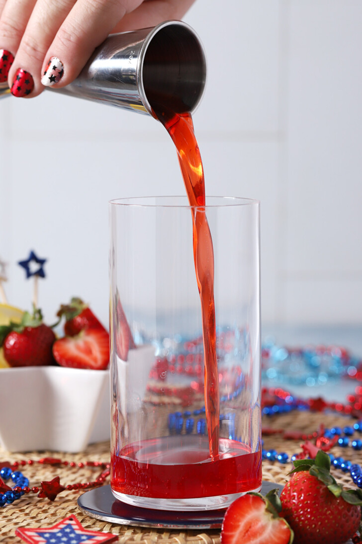 A hand pouring strawberry syrup from a jigger into a tall glass.
