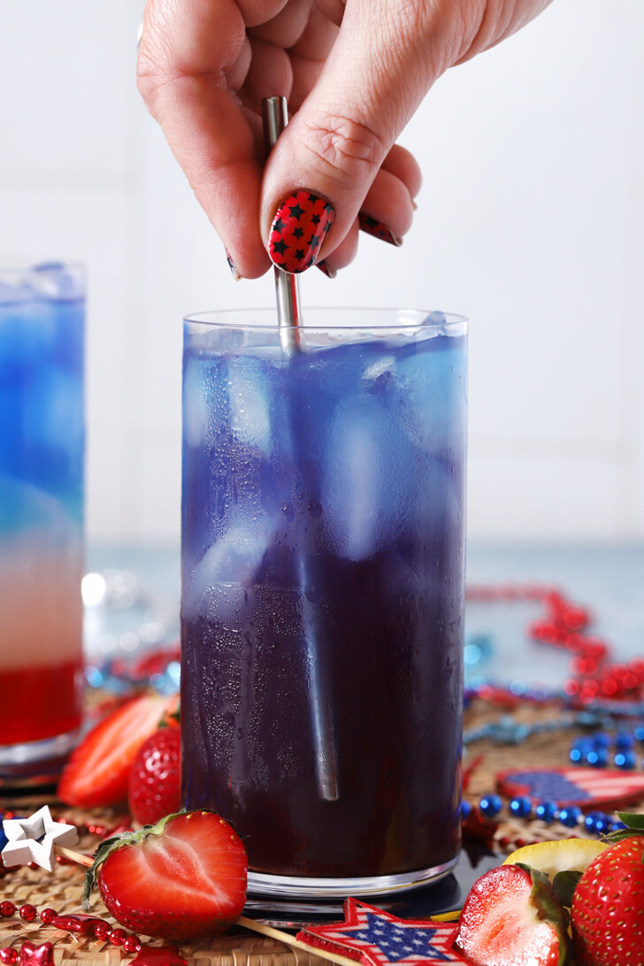 A hand stirring a red, white, and blue mocktail to combine the layers into a purple drink.