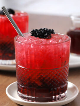 A glass of gin bramble on a coaster with a silver straw in it, garnished with a large blackberry.