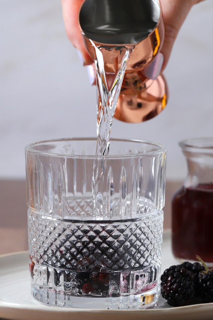 A hand pouring gin from a jigger into a rocks glass.