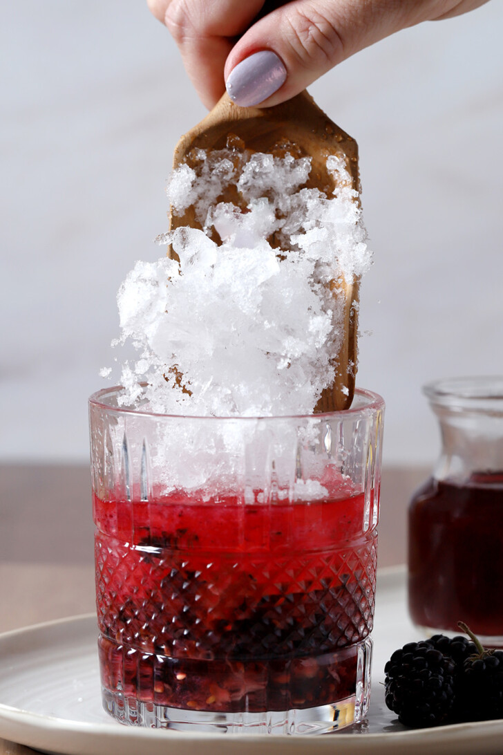 A hand pouring crushed ice from a jigger into a rocks glass.