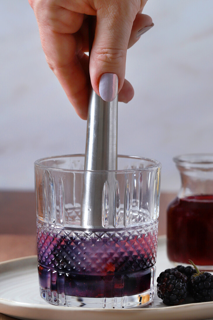 A hand using a muddler to crush blackberries in a rocks glass.