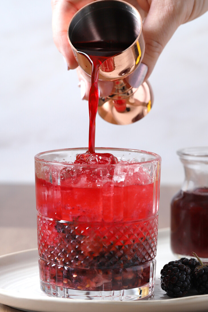 A hand pouring blackberry syrup from a jigger into a rocks glass filled with bramble cocktail.