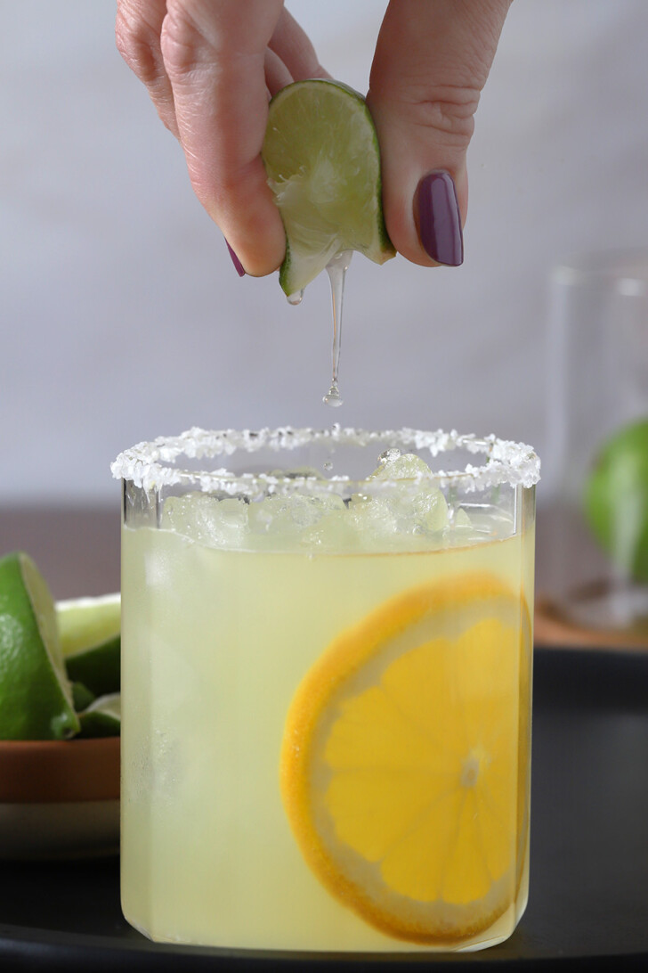 A hand squeezing a wedge of lime over a glass of orange margarita.