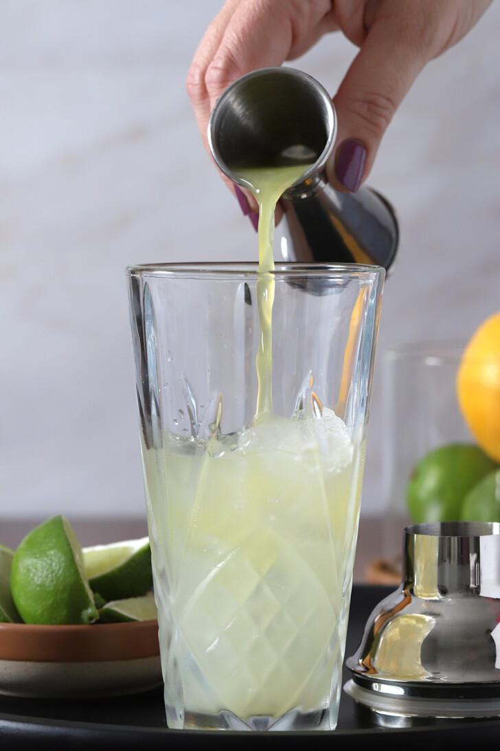 A hand pouring fresh orange juice from a jigger into a glass cocktail shaker.