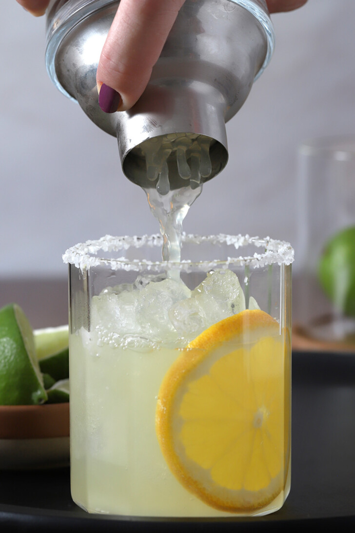 A hand holding a cocktail shaker upside down over a prepared glass filled with orange margarita.